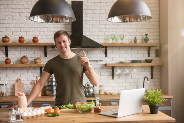 young smiling man standing in well-equipped kitchen looking for interesting ideas for breakfast checking recipe in his laptop , sunday morning - Foto, Imagem