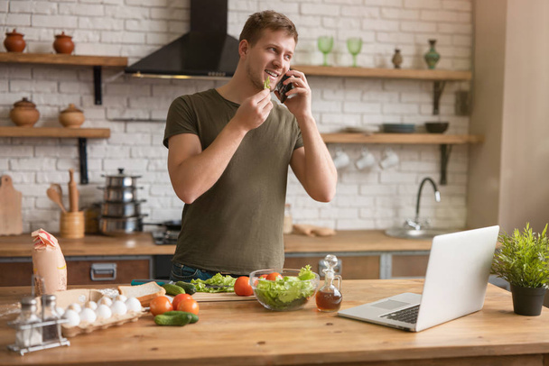young handsome man standing in well-equipped modern kitchen having pleasant phone conversation looking for interesting ideas for breakfast checking recipe in his laptop , sunday morning - Foto, Imagem