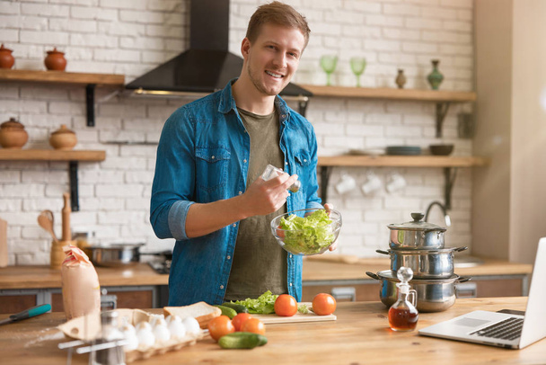 young handsome man adding salt and spices to salad while cooking lunch standing in well-equipped modern kitchen checking recipe in laptop , weekend vibes - Fotografie, Obrázek