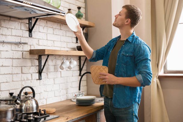 mladý pohledný muž stojící v dobře vybavené moderní kuchyni drží čistý talíř, zatímco utírá nádobí ručníkem po umytí, neděle ráno vibruje - Fotografie, Obrázek