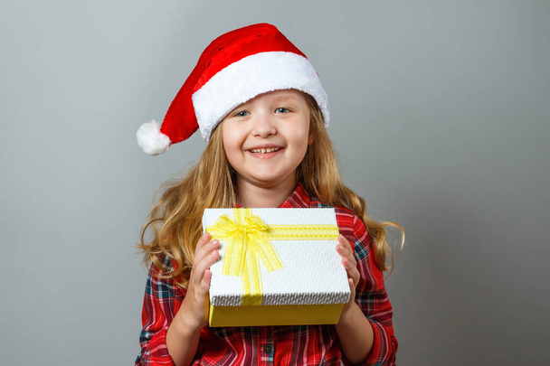 Christmas and New Year concept. Cute cheerful child holds a box with a gift in his hand. Charming little girl in santa hat and red dress on a gray background - Foto, Imagem