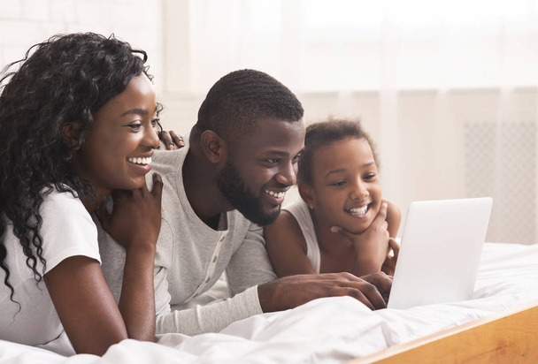 Família negra alegre de três usando laptop na cama juntos
 - Foto, Imagem