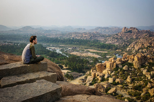 Um turista sentado à beira de uma colina. Bela vista das incríveis ruínas de Hampi da colina de Matanga. Karnataka, Índia
 - Foto, Imagem