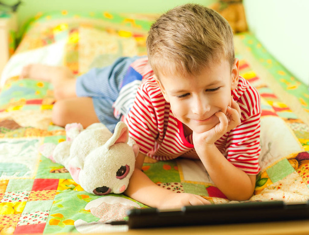 Niño preescolar acostado en la cama con la cara sonriente jugando juegos en el panel táctil
. - Foto, Imagen