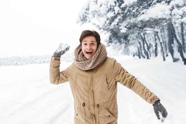 Счастливый молодой человек бросает снежок, глядя в камеру в лесу
 - Фото, изображение
