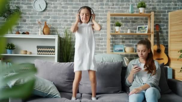 Little joyful girl in headphones jumping on sofa while mother using smartphone - Materiał filmowy, wideo