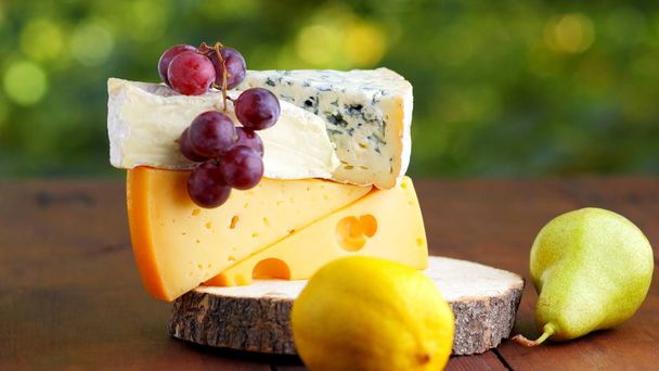 Camembert, brie, hard cheese and grapes on wooden stand. Pieces of different cheeses, pear and lemon on a blurred background. Dorblu and soft cheese on wooden boards. Widescreen - Foto, imagen