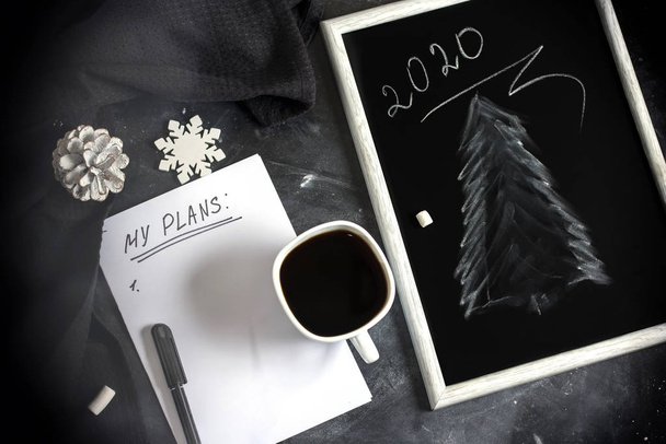 Χειμερινή Χριστουγεννιάτικη σύνθεση. Ένα φλιτζάνι καφέ σε ένα τραπέζι με ένα λευκό φύλλο χαρτί και ένα μαύρο πίνακα πλάκα με ένα σχέδιο ενός χριστουγεννιάτικου δέντρου. Χριστουγεννιάτικη διάθεση. Πάνω άποψη, επίπεδη lay - Φωτογραφία, εικόνα