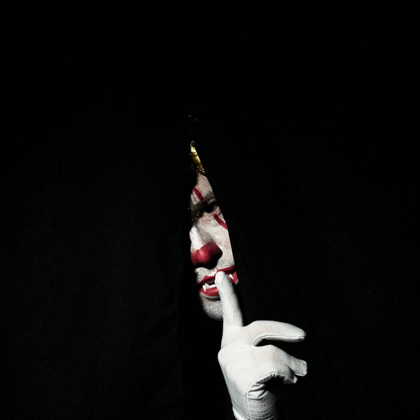 A terrible scary clown in a colored wig peeps out from behind black curtains. Shows a gesture of silence with a finger at the lips. - Фото, изображение