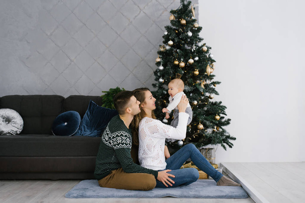 Μια ευτυχισμένη νεαρή οικογένεια με ένα μικρό γιο κάθεται στο πάτωμα κοντά σε ένα χριστουγεννιάτικο δέντρο στο σαλόνι - Φωτογραφία, εικόνα