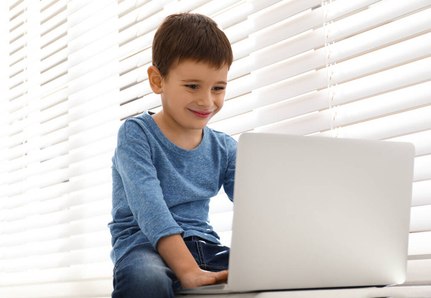 Happy little boy with modern laptop near window indoors - 写真・画像