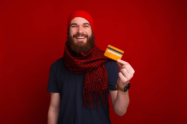 Νεαρός γενειοφόρος άνδρας χαμογελά και δείχνει μια πιστωτική κάρτα κοντά στο κόκκινο φόντο. - Φωτογραφία, εικόνα