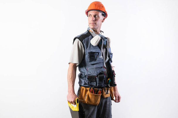 The builder in a helmet, and unloading, holds a hacksaw in his hands on a tree. On a light background. - Photo, Image