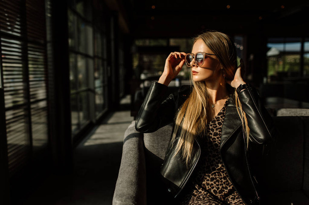 beautiful girl model in a restaurant in a beautiful loft interior. on the eyes of sunglasses, dressed in a leather black jacket, casual clothes. warm photography, sunlight - Photo, Image
