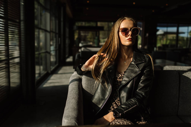 beautiful girl model in a restaurant in a beautiful loft interior. on the eyes of sunglasses, dressed in a leather black jacket, casual clothes. warm photography, sunlight - Foto, imagen