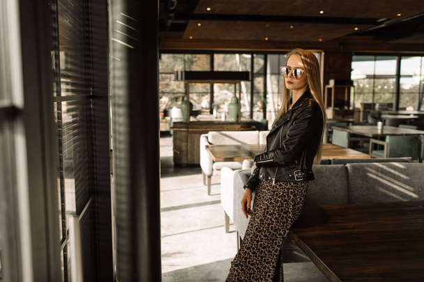 beautiful girl model in a restaurant in a beautiful loft interior. on the eyes of sunglasses, dressed in a leather black jacket, casual clothes. warm photography, sunlight - Фото, изображение