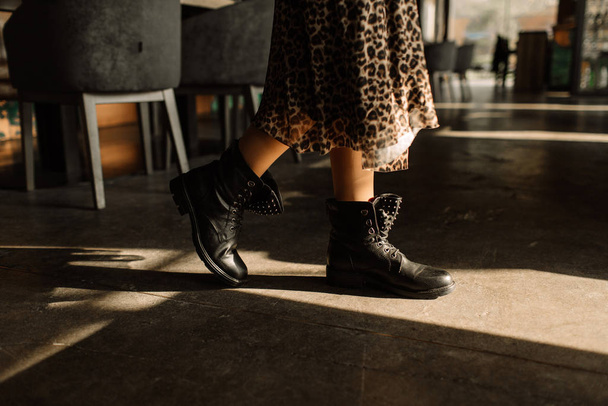 Girl in a leopard skirt and black boots in hard light on a concrete floor. Light key on boots - Photo, Image