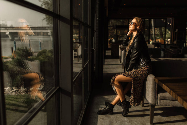 beautiful girl model in a restaurant in a beautiful loft interior. on the eyes of sunglasses, dressed in a leather black jacket, casual clothes. warm photography, sunlight - Photo, image
