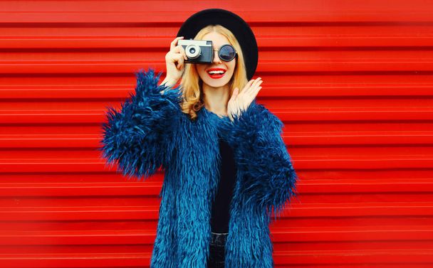 Portrait stylish surprised woman with retro camera taking picture wearing blue faux fur coat, round hat and sunglasses over colorful red wall background - Photo, image
