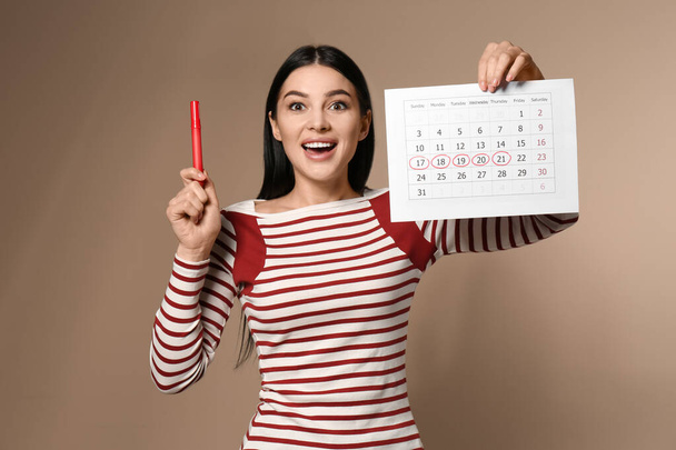 Young woman holding calendar with marked menstrual cycle days on beige background - Φωτογραφία, εικόνα