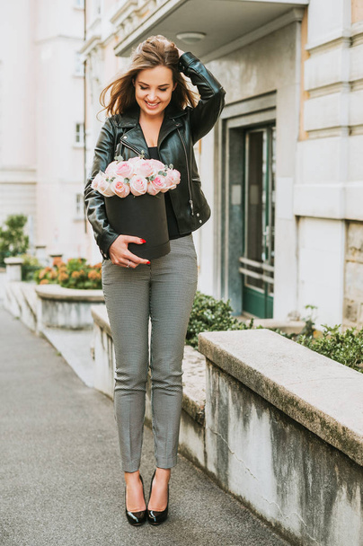 Portrait extérieur de jeune belle femme tenant boîte noire avec roses tendres roses, veste en cuir et pantalon à carreaux
 - Photo, image