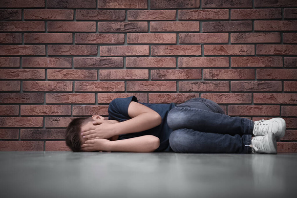 Sad little boy closing eyes with hands on floor near brick wall. Child in danger - Foto, immagini