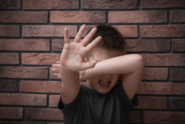 Scared little boy closing eyes with hand near brick wall. Child in danger - Photo, Image