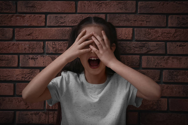 Scared little girl closing eyes with hands near brick wall. Child in danger - Photo, Image