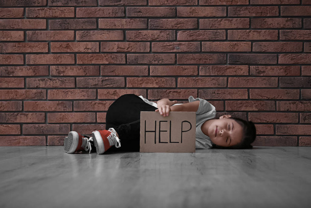 Sad little girl with sign HELP lying on floor near brick wall. Child in danger - Foto, immagini