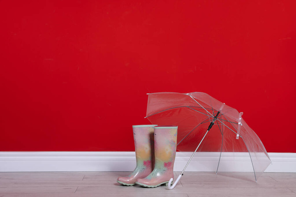 Beautiful transparent umbrella and rubber boots near red wall. Space for text - Zdjęcie, obraz