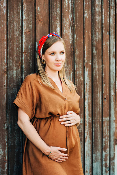 Outdoor portrait of stylish pregnant woman, wearing brown dress and stylish headband - Photo, Image