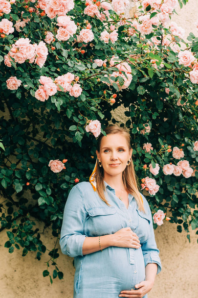 Retrato al aire libre de una mujer embarazada con estilo posando en un jardín de rosas, usando vestido de mezclilla
 - Foto, Imagen