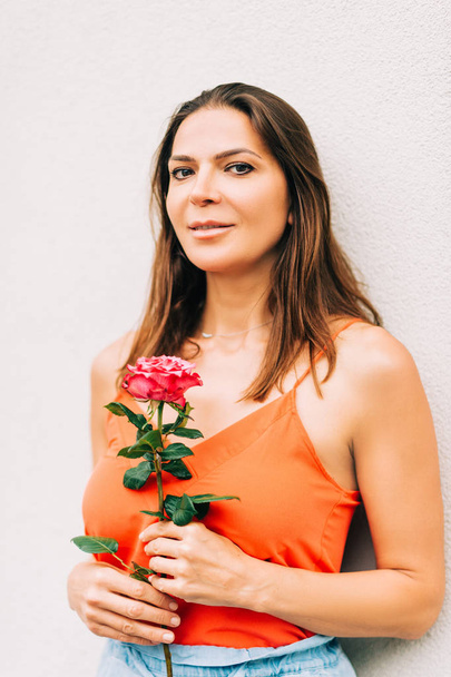 Portret atrakcyjnej kobiety trzymającej różowy kwiat, noszącej pomarańczowe ubrania - Zdjęcie, obraz
