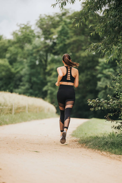 Portrait of young beautiful fit woman jogging in nature, wearing black activewear, athlete model posing outdoors, sport fashion, back view - 写真・画像