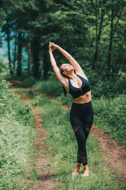 Fiatal nő gyakorló jóga a nyári erdőben, visel fekete leggings és top - Fotó, kép