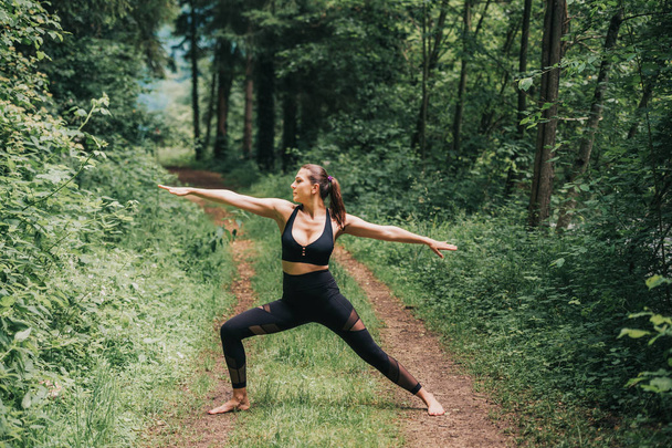 Молодая женщина занимается йогой в летнем лесу, носит черные леггинсы и топ
 - Фото, изображение