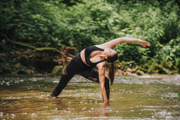 Mujer joven en forma practicando yoga, de pie en el río, con leggins negros y parte superior
 - Foto, imagen