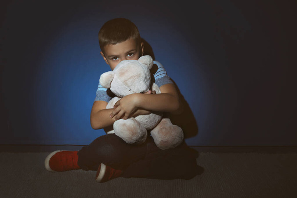 Sad little boy with teddy bear near blue wall. Domestic violence - 写真・画像