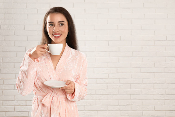 Young woman in bathrobe with cup of coffee near white brick wall - Φωτογραφία, εικόνα