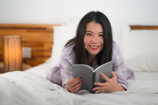 young beautiful and sweet Asian Korean woman in bed reading book novel feeling happy and playful or studying at home wearing cute pajamas looking cozy and homey - Foto, Imagen