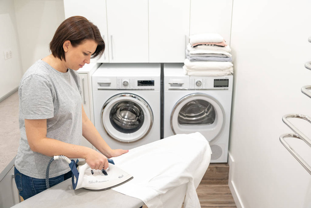 Mädchen in der Waschküche bügelt weißes Hemd an Bord mit Waschmaschine im Hintergrund - Foto, Bild