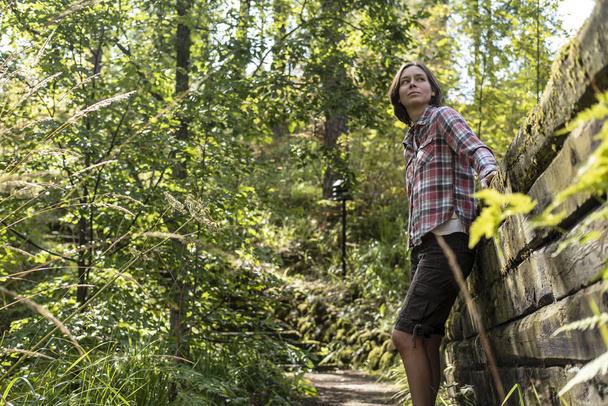 Jolie femme se tient sur un sentier forestier, près d'un vieux mur de rondins et regarde loin, au milieu d'une belle forêt, dans un parc un jour d'été
. - Photo, image