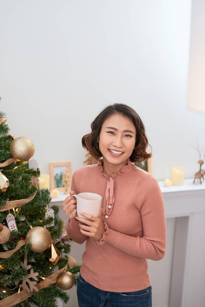 Gyönyörű lány göndör haj kezében egy csésze kávét a háttérben a karácsonyfa - Fotó, kép