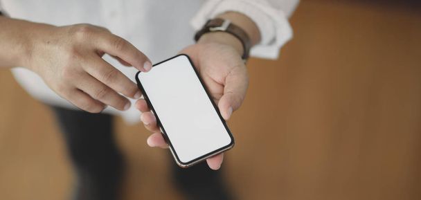 Vista de cerca del hombre usando el teléfono inteligente de pantalla en blanco mientras está de pie en su habitación de la oficina
  - Foto, Imagen
