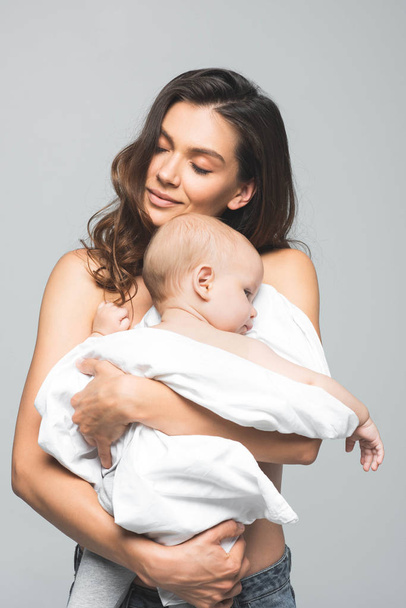 Porträt einer glücklichen nackten Mutter mit geschlossenen Augen, die ihr Baby umarmt, isoliert auf grau - Foto, Bild