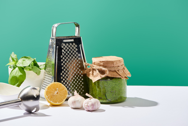 соус песто в банке рядом с ингредиентами, блендер и терка на белом столе изолированы на зеленый
 - Фото, изображение
