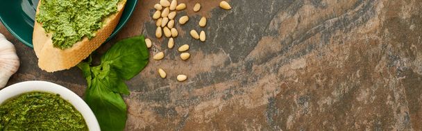 bovenaanzicht van stokbrood plak met pesto saus op bord in de buurt van verse ingrediënten op stenen oppervlak, panoramisch schot - Foto, afbeelding