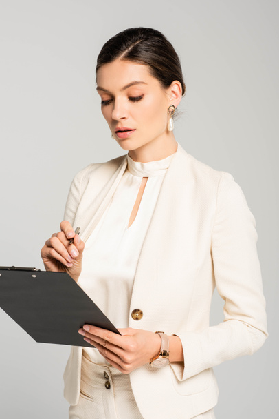 beautiful businesswoman in white suit writing in clipboard, isolated on grey - Фото, изображение