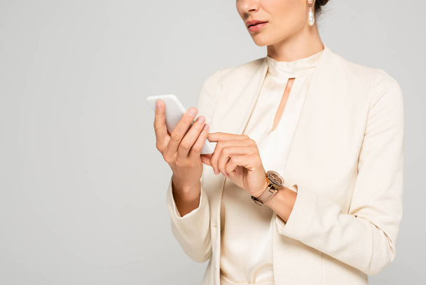 bijgesneden weergave van zakenvrouw in wit pak met behulp van smartphone, geïsoleerd op grijs - Foto, afbeelding