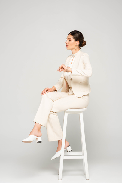 elegant businesswoman in white suit sitting on stool, on grey - Фото, изображение